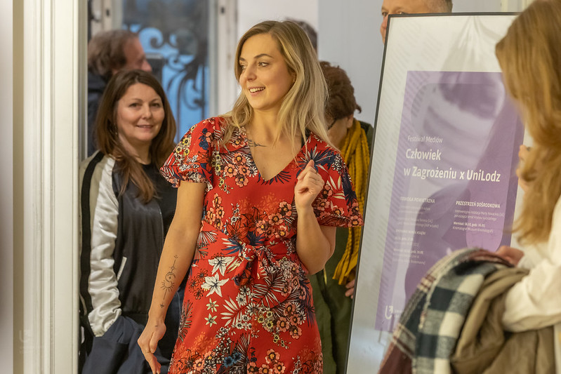 Marta Nowacka w sali wystawy, obok stoi plakat z informacją o festiwalu.